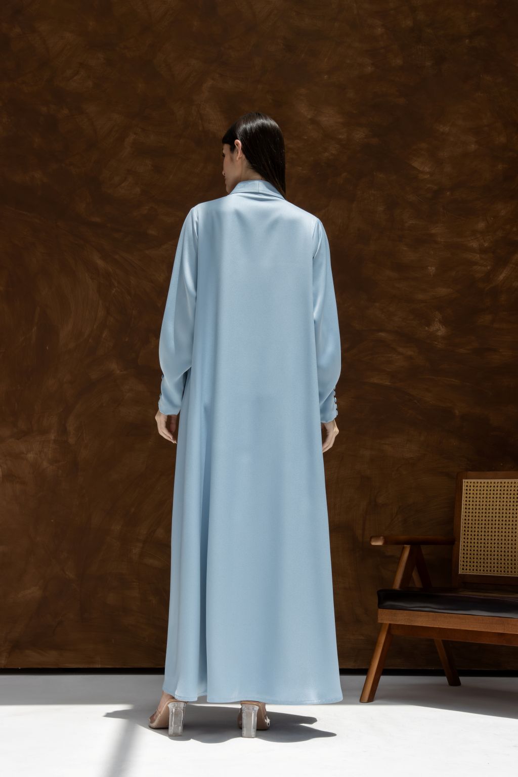 Back view of woman wearing elegant Buttoned Breeze blue abaya by Selected Lines standing against brown wall