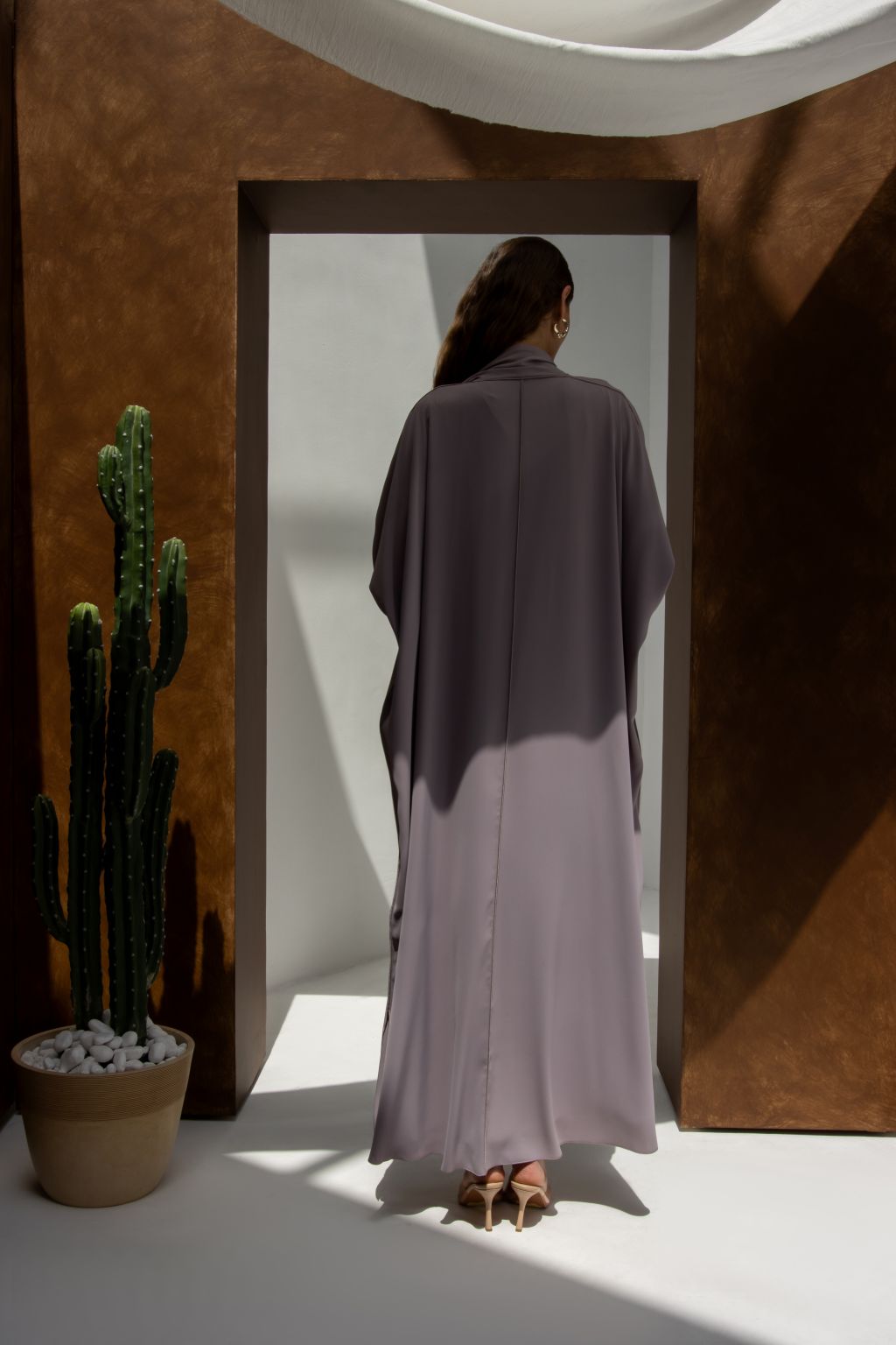 Back view of a model wearing elegant signature abaya in soft gray from Selected Lines standing by a cactus plant