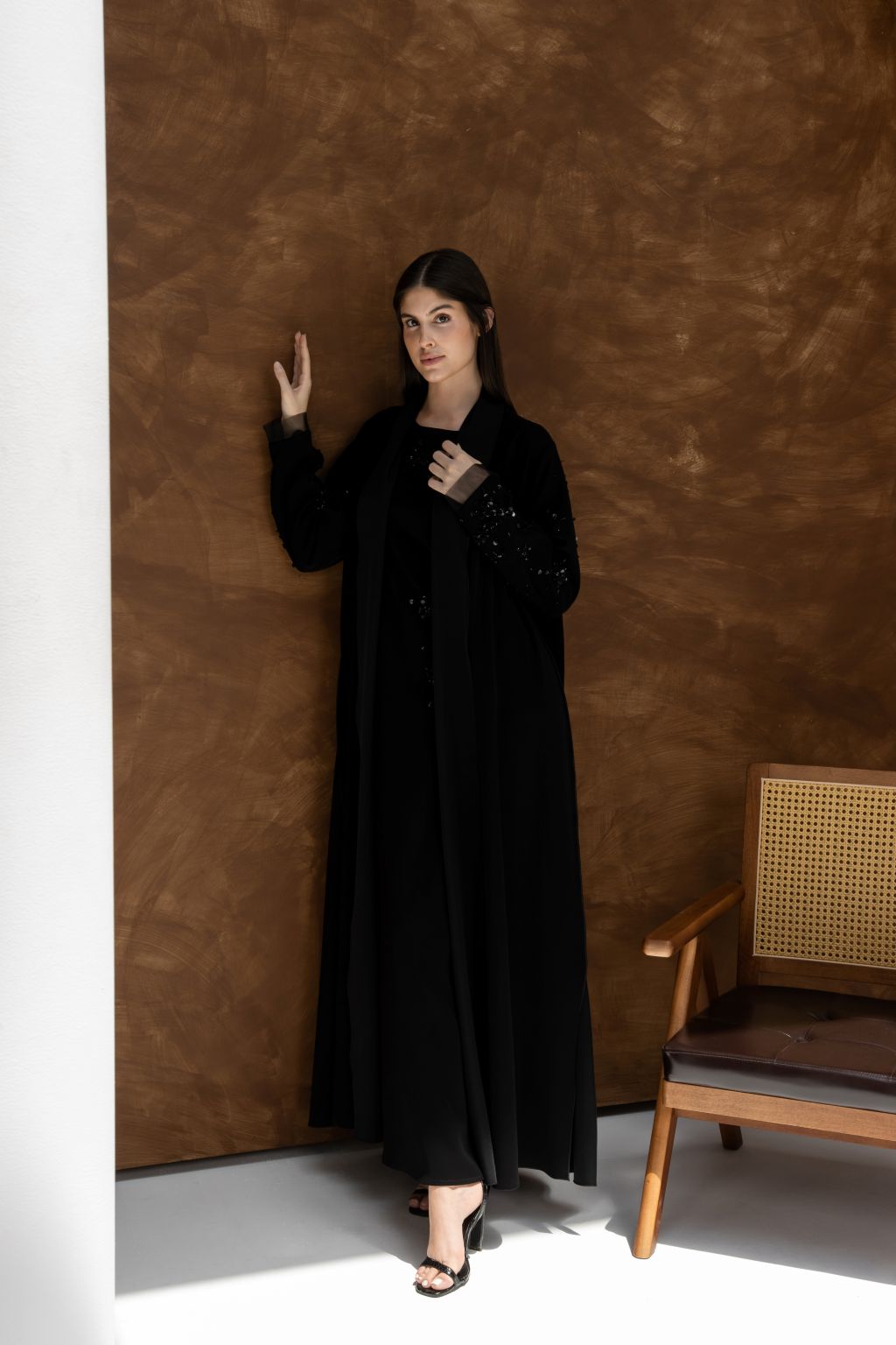 Woman wearing black La Lueur abaya Set from Selected Lines standing by a brown wall next to a wooden chair