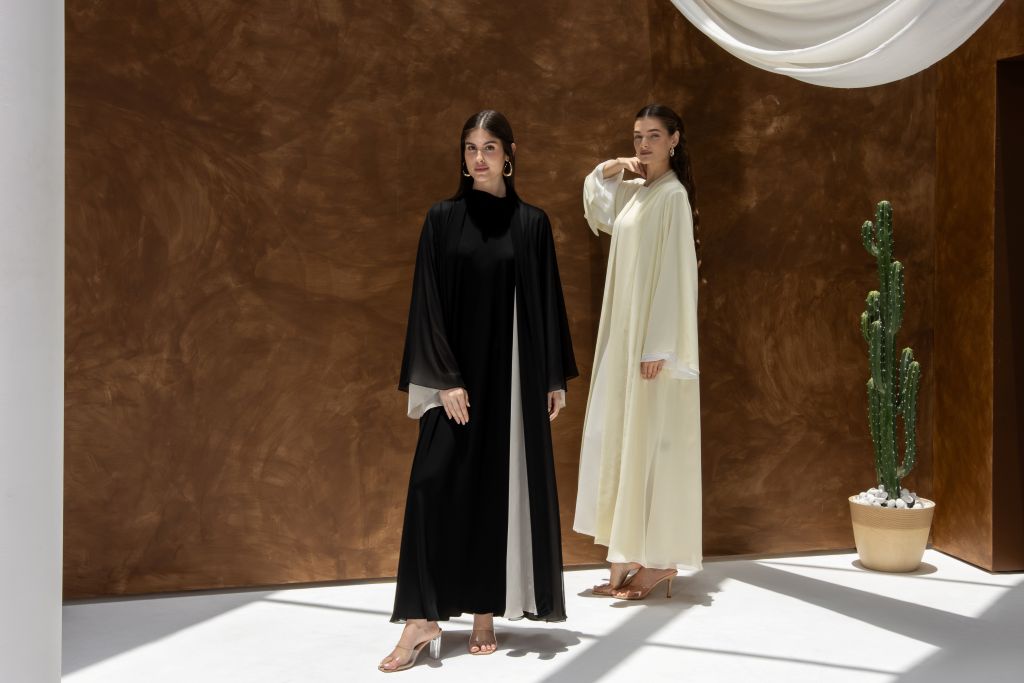 Two women wearing Selected Lines Layered Grace Abaya in black and cream standing against brown wall with sunlight