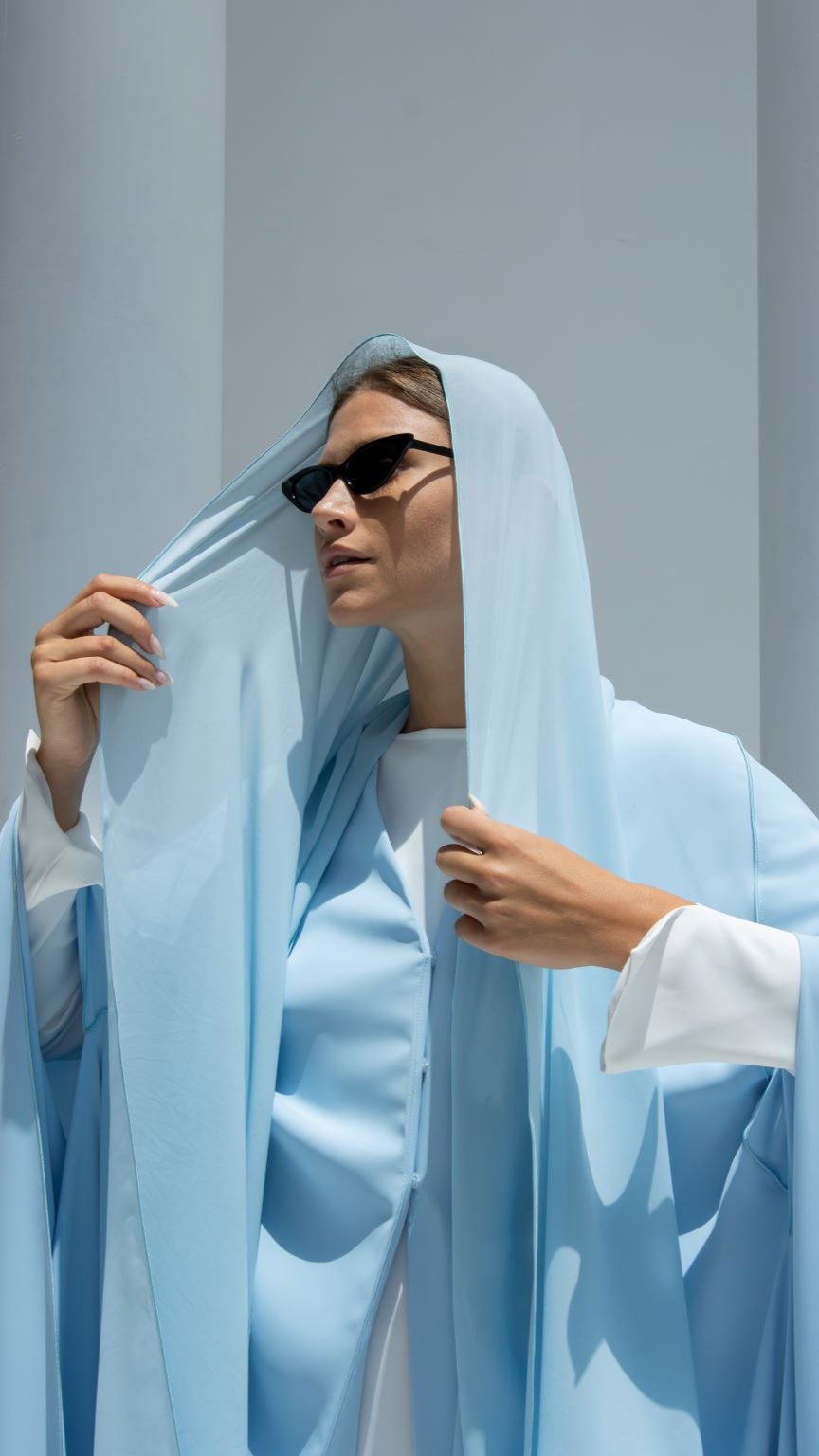 Elegant luxury Noura Abaya in soft blue draped over a stylish model with sunglasses