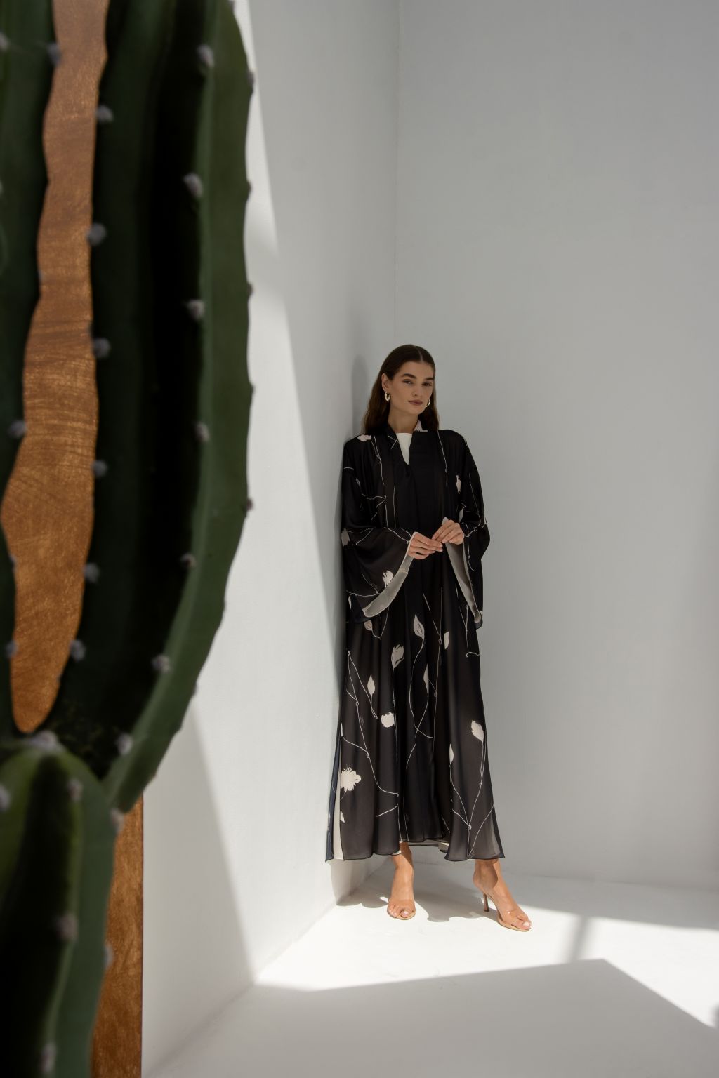 Selected Lines signature leaves black abaya worn by model standing in minimal white room with cactus plant
