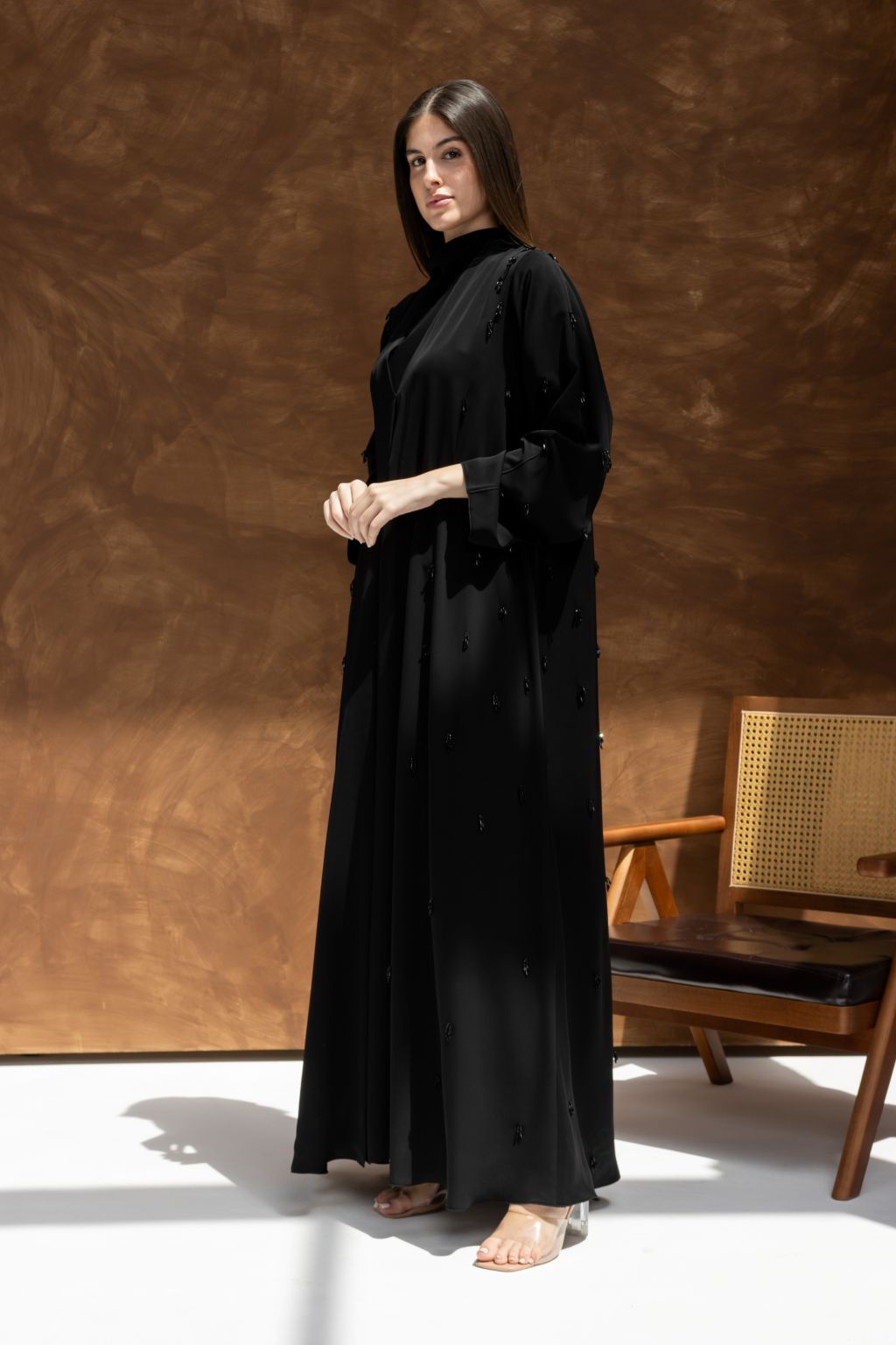 Woman wearing a timeless black abaya gown by Selected Lines standing next to a wooden chair
